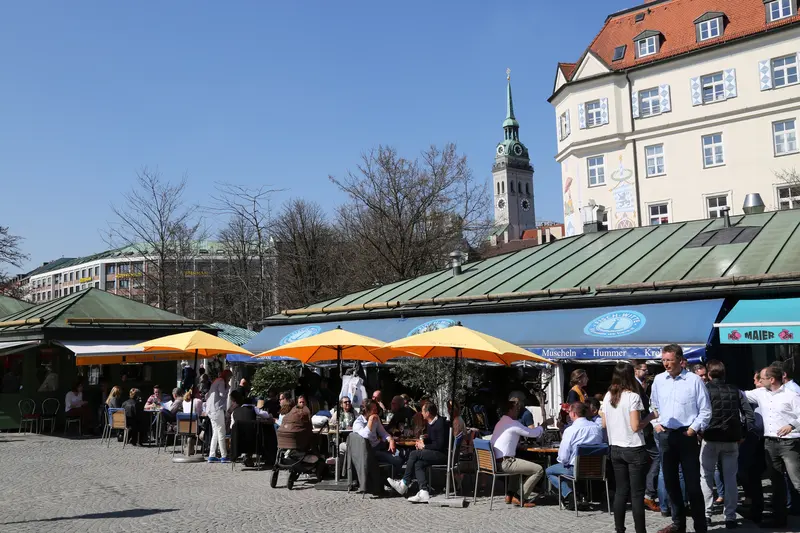 Market stalls with orange umbrellas and seated crowds at the Viktualienmarkt in Munich, with the St. Peter's church tower behind